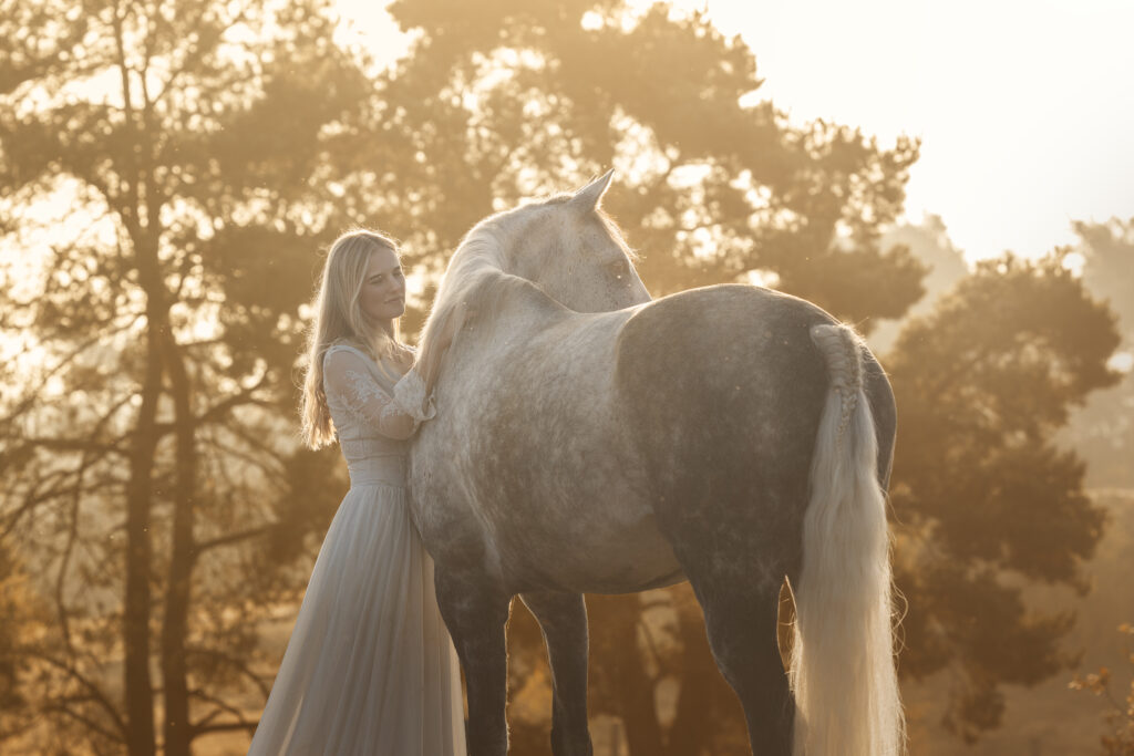 Mädchen in weißem Kleid mit ihrem Apfelschimmel in der Heide im Winter im Sonnenaufgang