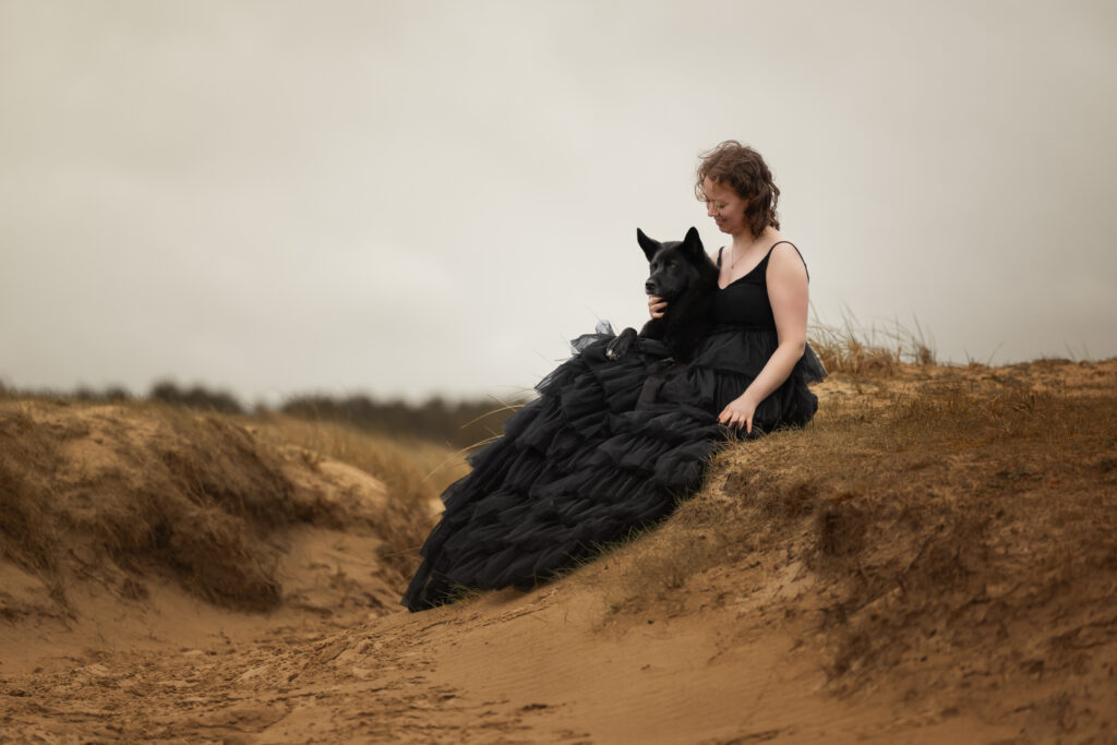 Junge Frau in schwarzem Kleid sitzt mit schwarzem Hund in den Dünen